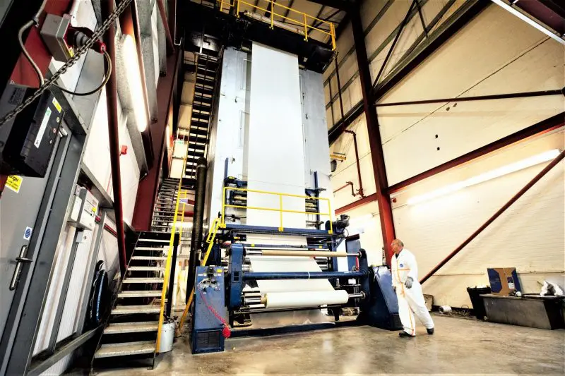 Prepreg-Retouched-2-800x533 A worker inspects a large industrial paper machine inside a factory with metal stairs and railings nearby.