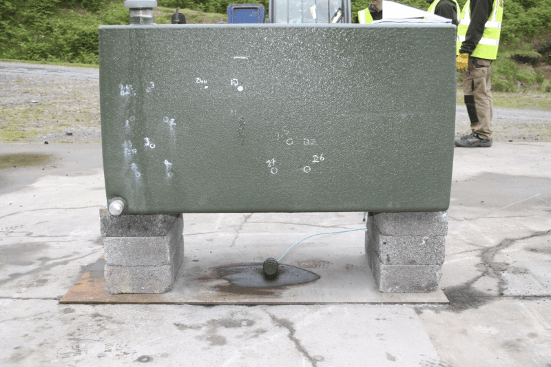 Green metal tank on breeze blocks, leaking liquid onto a concrete slab outdoors.