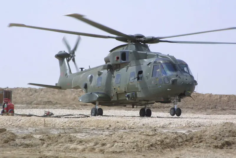 About-Merlin-Desert-800x534 Green military helicopter landed on a sandy area with equipment and cables nearby under a clear sky.