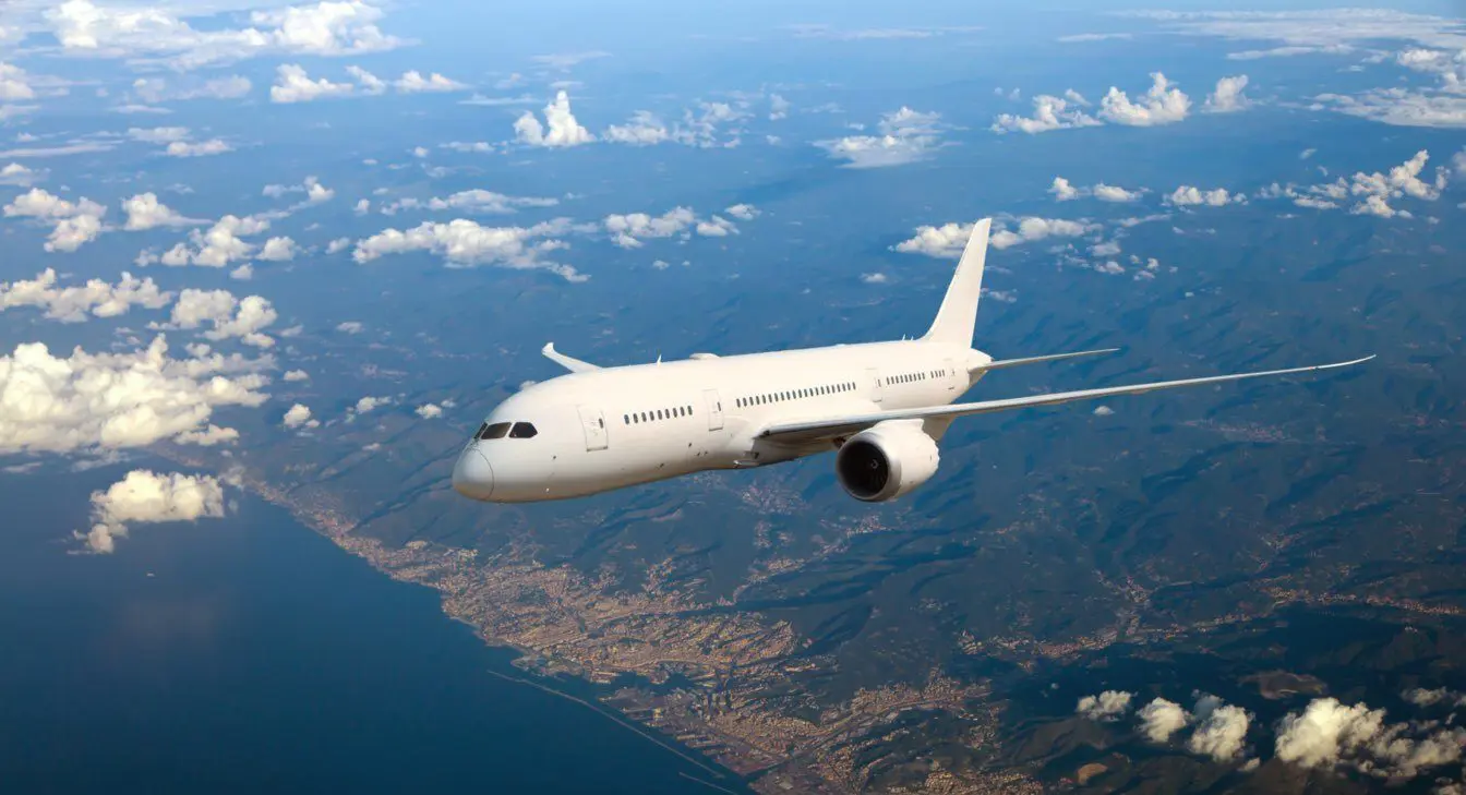 Boeing-787-dreamliner-1-e1590681938108 A white airplane flying above the clouds with land and coastline visible below.