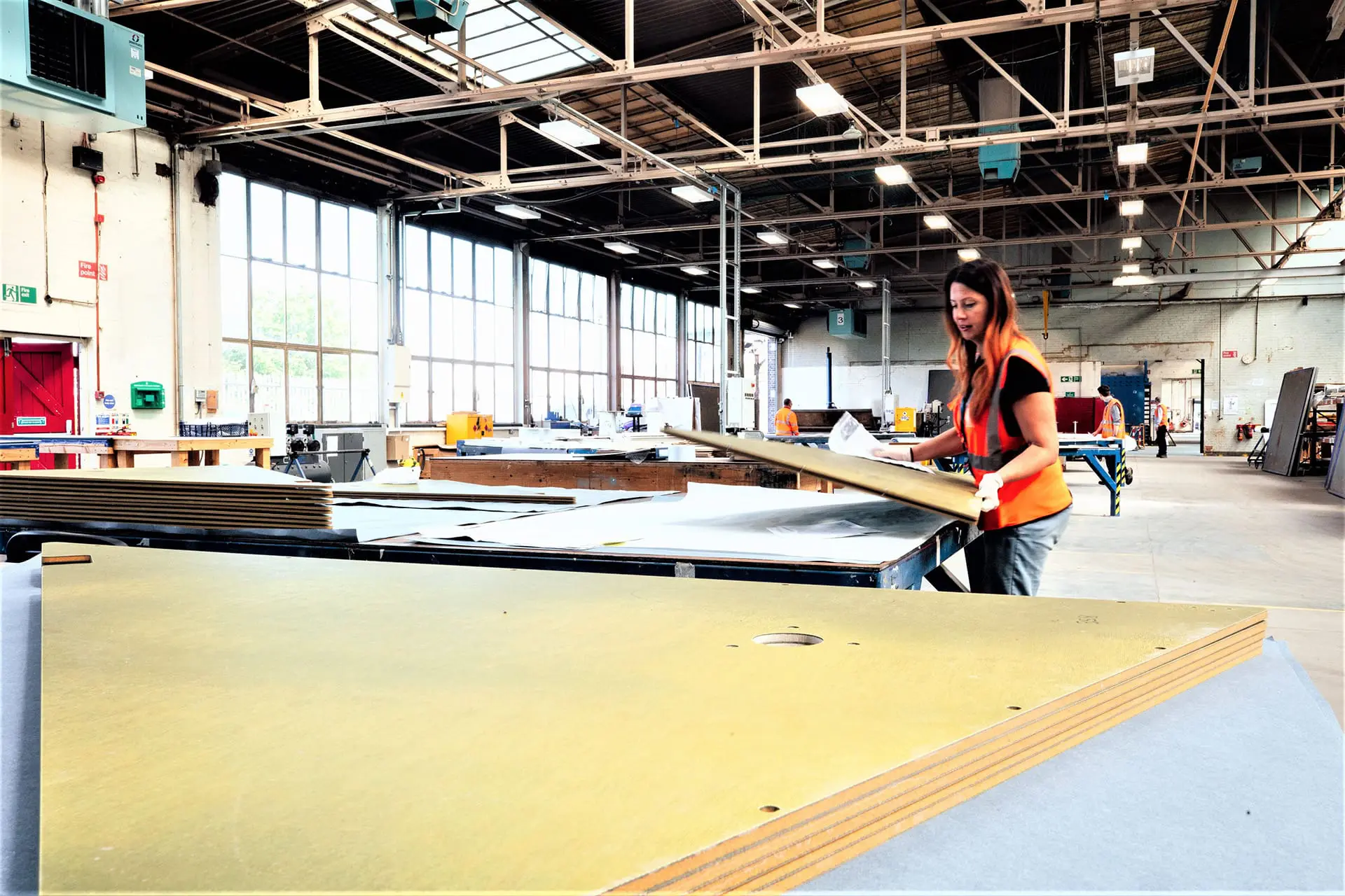 Bonding-and-Assembly-Retouched A woman in a workshop moves a large sheet of material on a table in a bright, spacious industrial space.