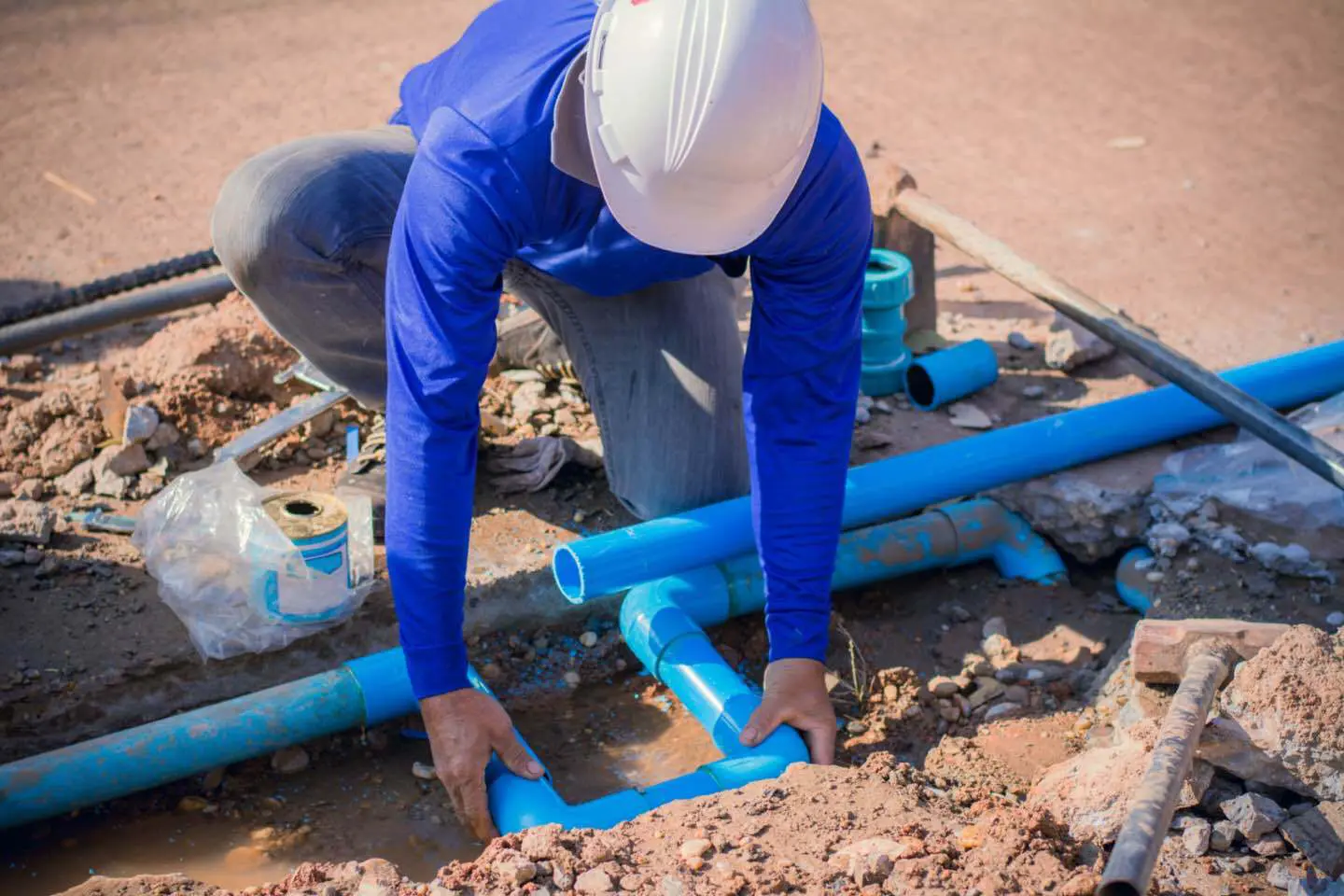 Construction-header-image-domestic-drainage-1 Worker in a blue shirt and white helmet installing blue PVC pipes in a muddy construction site.