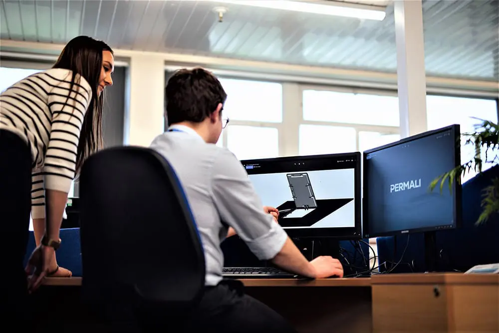 Design-Consultancy-Retouched Two people look at a 3D design on a computer in an office with dual monitors.