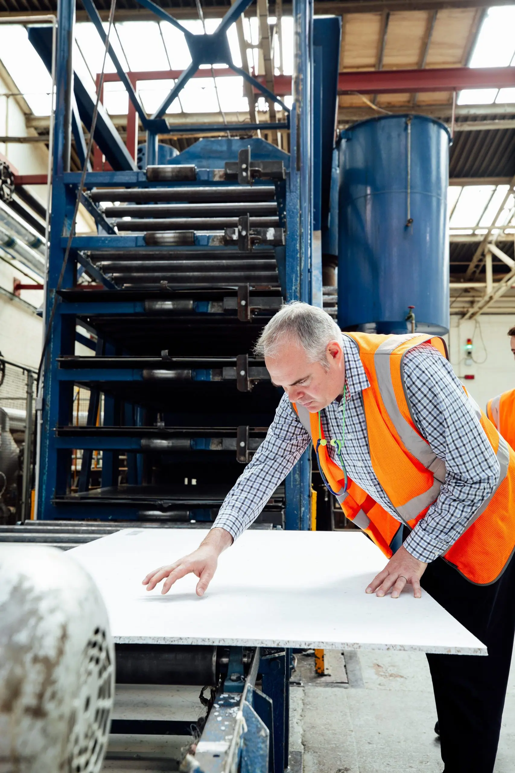 Permali Shoot Proofs-038 Man in an orange safety vest inspecting a large white sheet in a factory with industrial equipment.