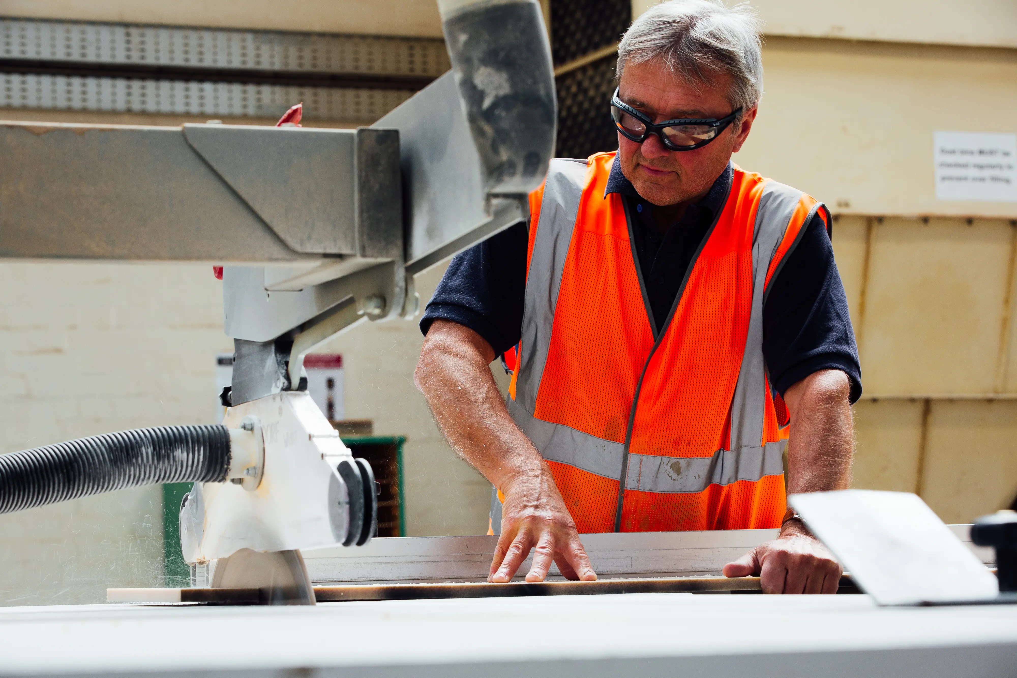 Man in an orange safety vest operates a table saw, cutting wood in a workshop.