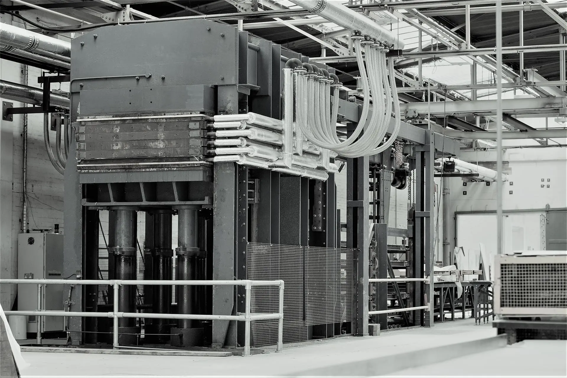 Press Large industrial machine with multiple pipes and cables in a factory setting, surrounded by metal railings.