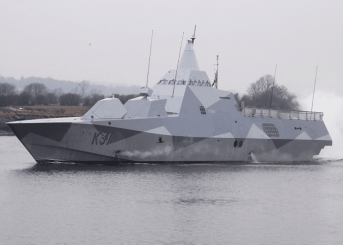 A modern navy warship with a camouflage pattern sails on calm water near a shoreline with trees.