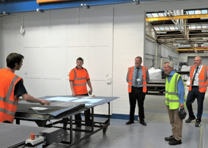 Five men in safety vests stand and watch a worker handling materials in an industrial warehouse.