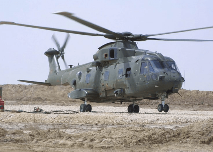 A military helicopter with twin rotors sits on rocky ground in a desert-like area under a clear sky.