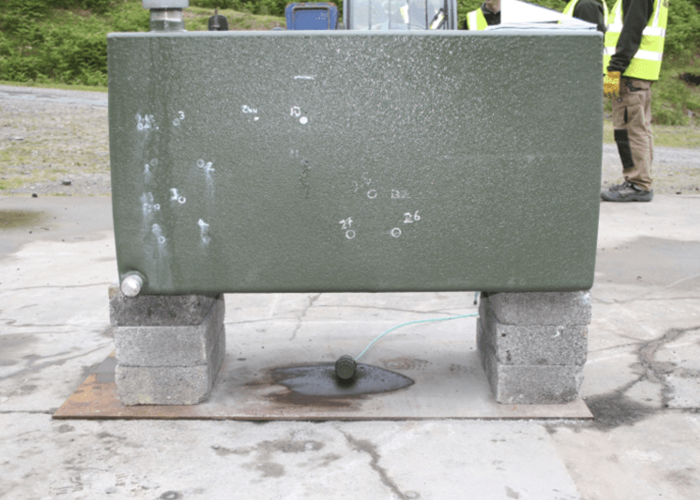 Battlejacket-Case-Study-2-800×533 (1) A green metal tank on concrete blocks with bullet holes and a liquid leaking onto the ground.