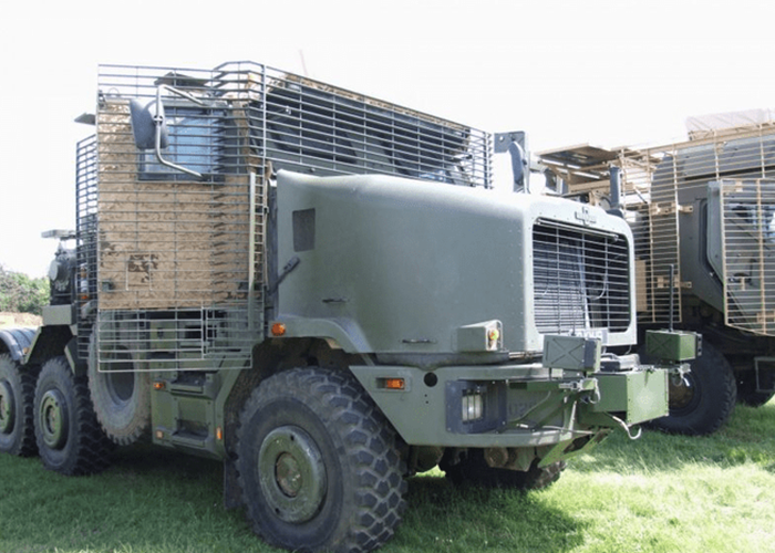 HETatDvD-800×600 Military truck with protective cage armor parked on grass in daylight.