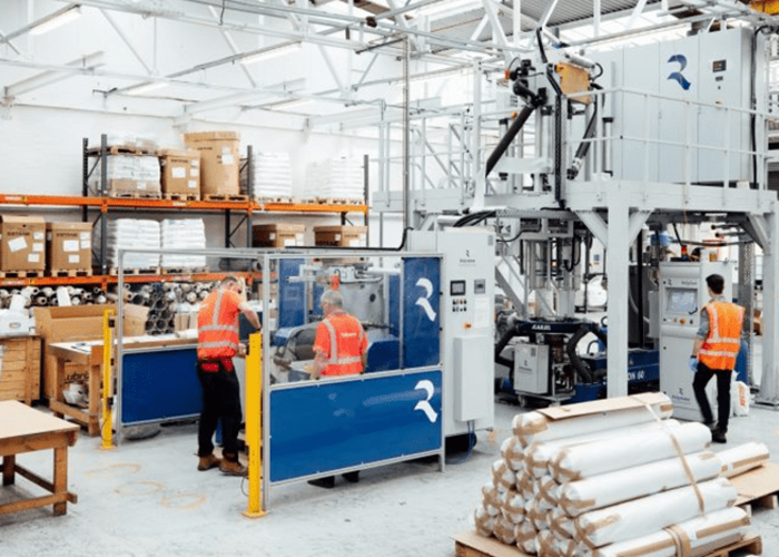 Three workers in safety vests operate machinery in a bright, organized industrial warehouse with stacked supplies.