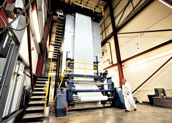 A worker stands by a large industrial paper printing press inside a high-ceilinged factory.