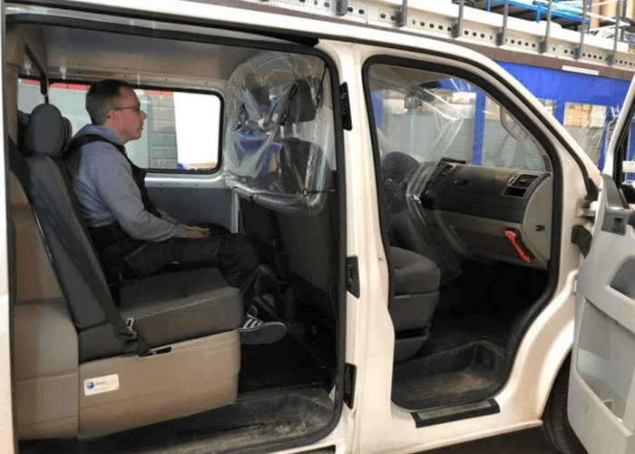 A man sits alone in the back seat of a white van with a plastic divider behind the front seats.