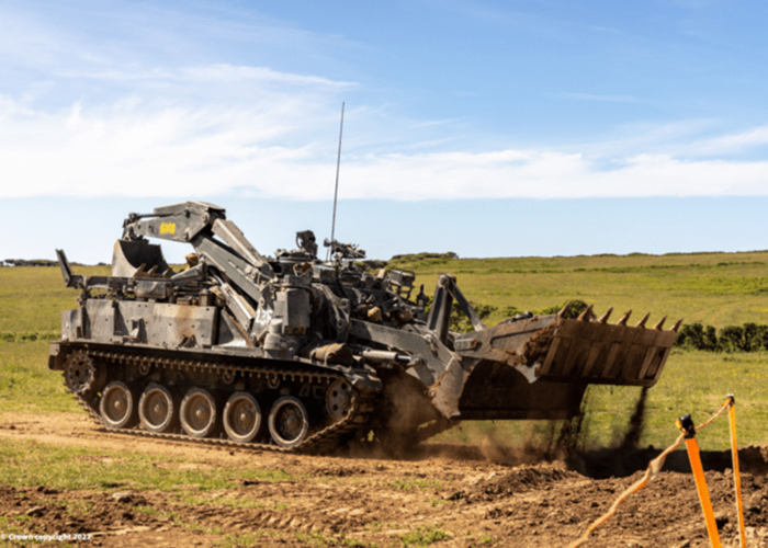 A large military tracked vehicle with a front loader moves dirt on a grassy field under a blue sky.