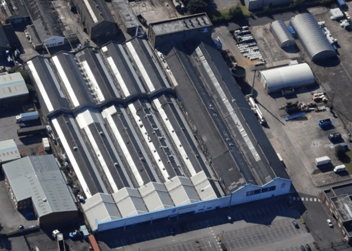 Aerial view of a large industrial warehouse with a zigzag roof and adjacent buildings in a paved area.