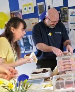 Two people serve baked goods at a table with cakes and pastries, in a room decorated with balloons.