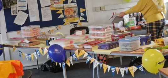 A table with cakes, drinks, balloons, and bunting set up for a bake sale in a decorated room.