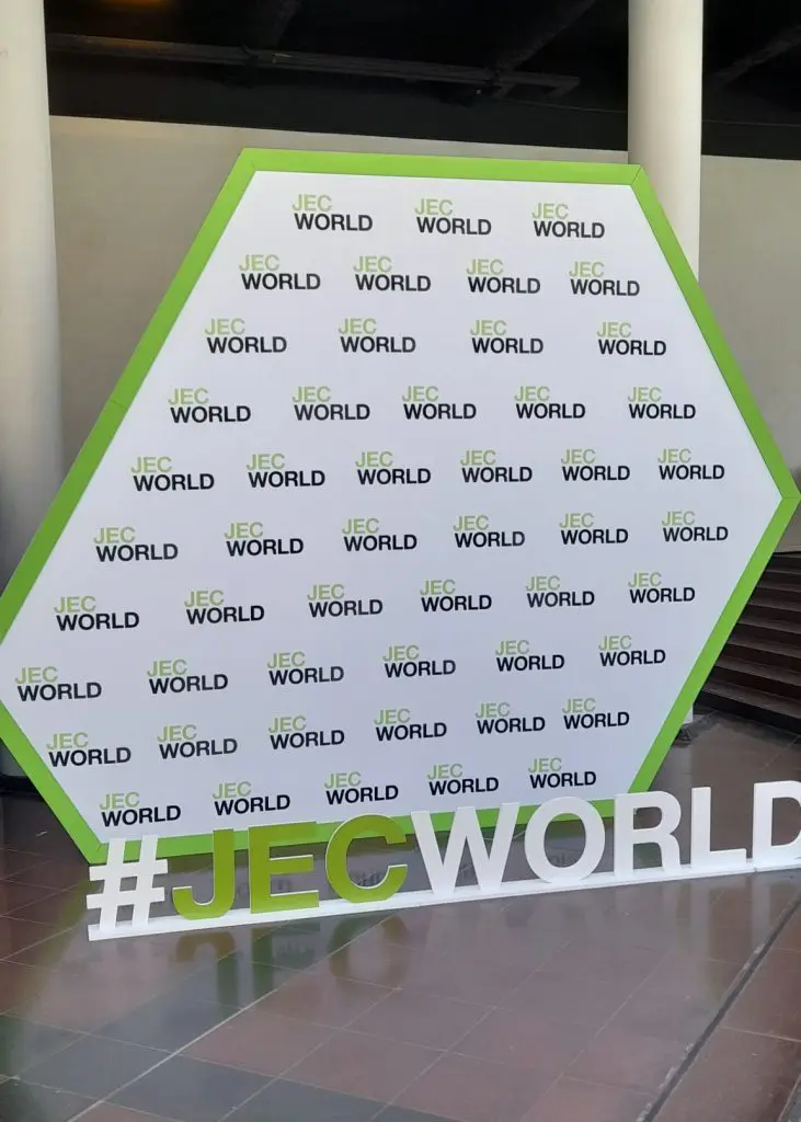 A hexagonal JEC World display with a #JECWORLD hashtag in front, set indoors near stairs.