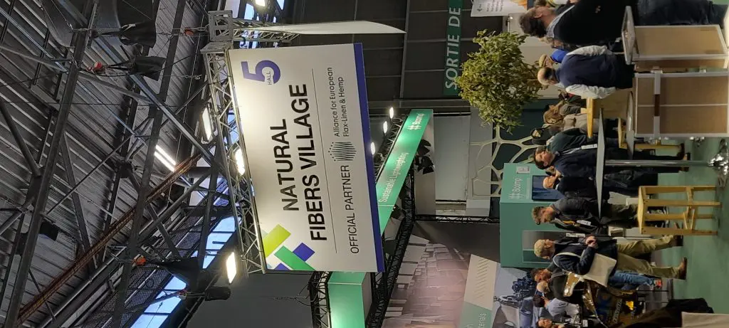 JEC-2025-2 People walk under a large Natural Fibers Village sign at an indoor exhibition with booths and displays.