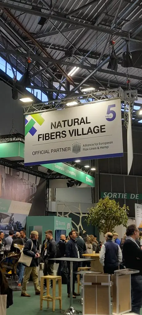 People walk under a large Natural Fibers Village sign at an indoor exhibition with booths and displays.
