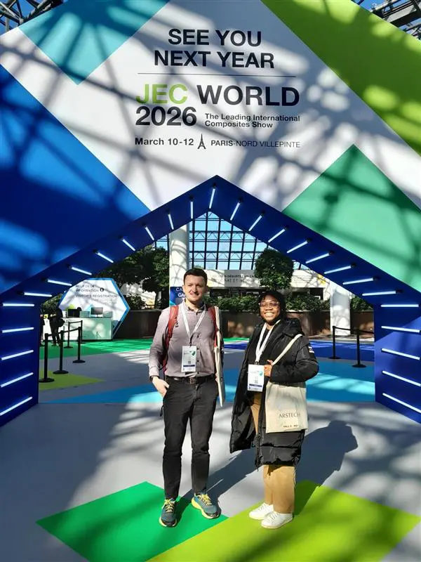 Two people stand smiling under a sign for JEC World 2026 at a bright, modern event venue.