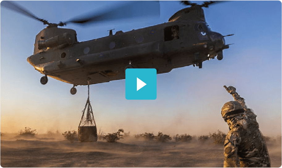 A soldier signals as a military helicopter lifts cargo in a dusty outdoor setting at sunset.
