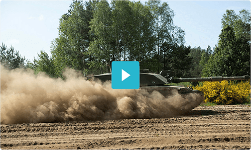 A tank driving through dirt, kicking up dust, with trees and greenery in the background.