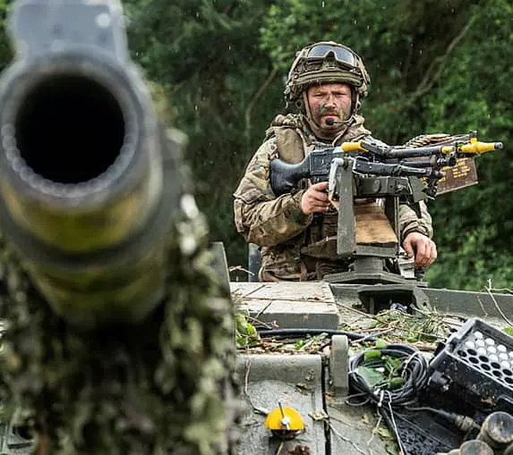 land-defence-9-8 A soldier in camouflage gear sits atop a military vehicle, aiming a machine gun, with a blurred barrel in the foreground.