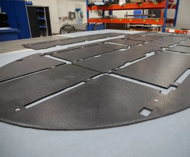 Several black, flat, rectangular panels with slots lay on a table in an industrial workshop.