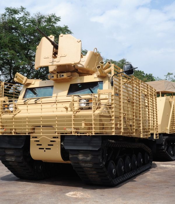 Warthog_TCV_First-Article Tan-colored armored military vehicle with tracks and a mounted gun, parked on a paved area outdoors.