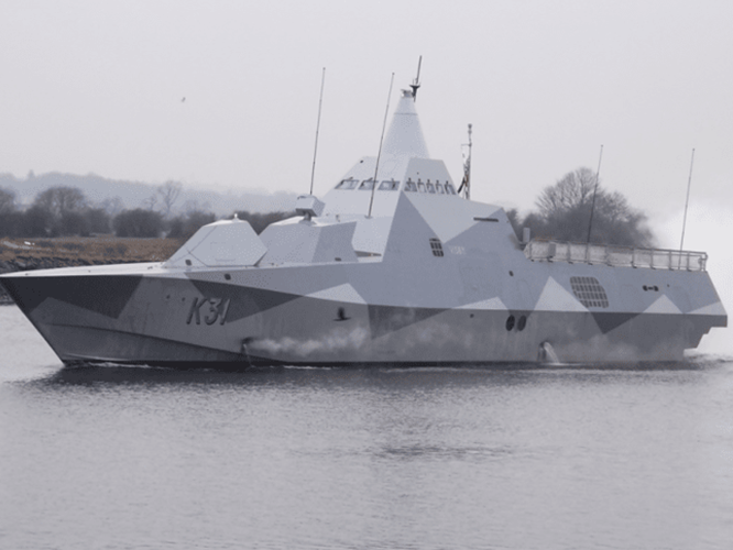 1-1-800×534 A modern navy warship with a camouflage pattern sails on calm water near a shoreline with trees.