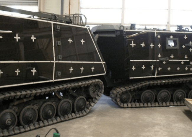 3.png-800×432 Two black, tracked armored vehicles with white outlined crosses parked indoors, facing each other.