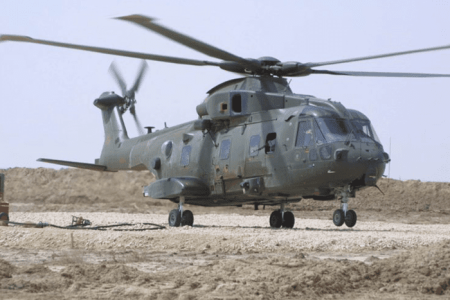About-Merlin-Desert-800x534 A military helicopter with twin rotors sits on rocky ground in a desert-like area under a clear sky.