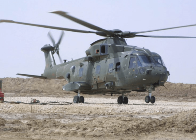 About-Merlin-Desert-800×534 A military helicopter with twin rotors sits on rocky ground in a desert-like area under a clear sky.