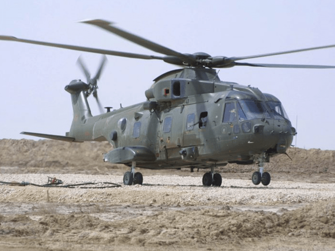 About-Merlin-Desert-800×534 A military helicopter with twin rotors sits on rocky ground in a desert-like area under a clear sky.