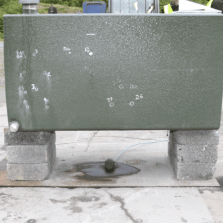 Battlejacket-Case-Study-2-800x533 (1) A green metal tank on concrete blocks with bullet holes and a liquid leaking onto the ground.