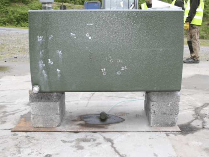 Battlejacket-Case-Study-2-800×533 (1) A green metal tank on concrete blocks with bullet holes and a liquid leaking onto the ground.