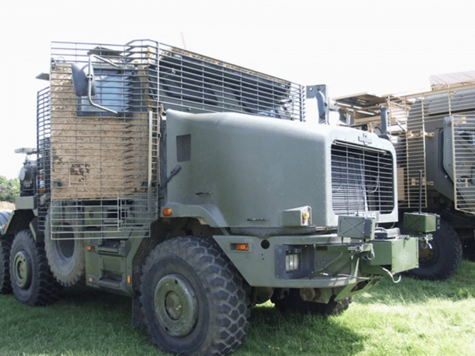 HETatDvD-800×600 Military truck with protective cage armor parked on grass in daylight.