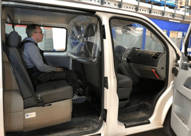 Screen-3-800×600 A man sits alone in the back seat of a white van with a plastic divider behind the front seats.