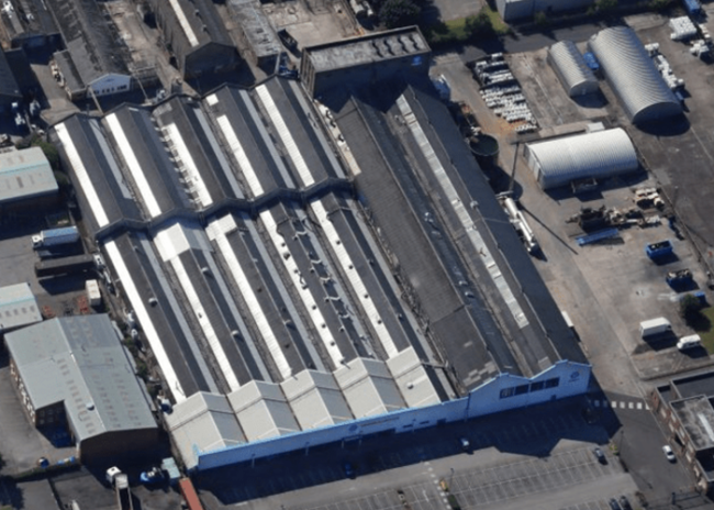 Aerial view of a large industrial warehouse with a zigzag roof and adjacent buildings in a paved area.