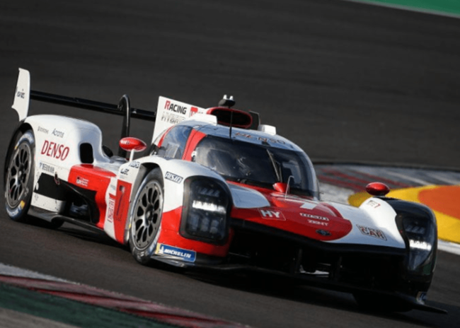 Red and white Toyota race car speeds around a track corner during a motorsport event.