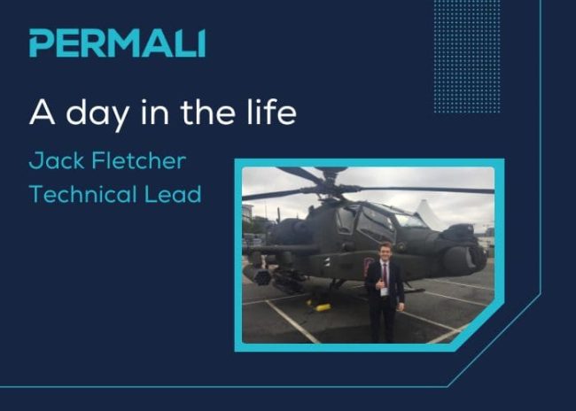 Permali Blog Feature Image Size A man in a suit stands in front of a military helicopter in a parking lot, smiling at the camera.