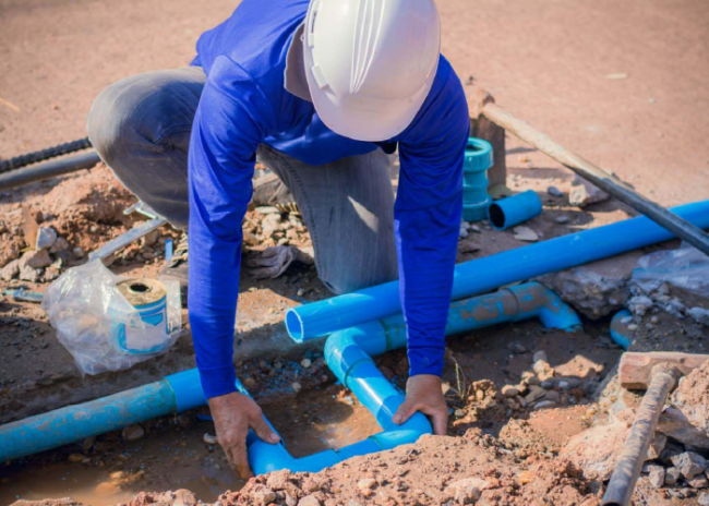 Permali_Applications-Polyurea-Coatings_Featured-Image Worker in a blue shirt and hard hat installing blue PVC pipes in a muddy construction site.