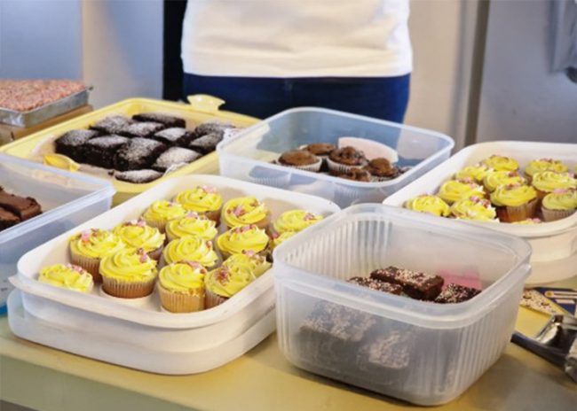 Various trays and containers of cupcakes and brownies on a table, with a person standing behind them.