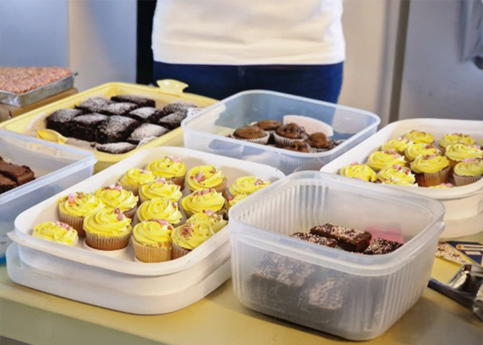 Various trays and containers of cupcakes and brownies on a table, with a person standing behind them.
