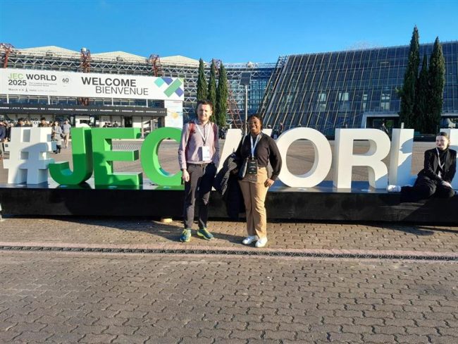 JEC-2025-4 Two people stand in front of a large #JECWORLD sign outside a convention center on a sunny day.