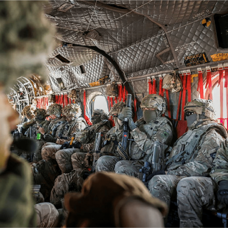 Soldiers in camouflage gear and helmets sit inside a military aircraft, holding rifles and facing forward.