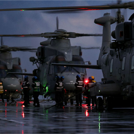 Navy personnel work on helicopters at night, with red and green lights reflecting on the wet ground.