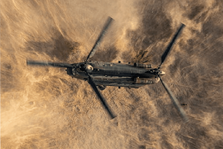 Image-1 A military helicopter flies over a sandy landscape, kicking up a cloud of dust beneath its spinning rotors.